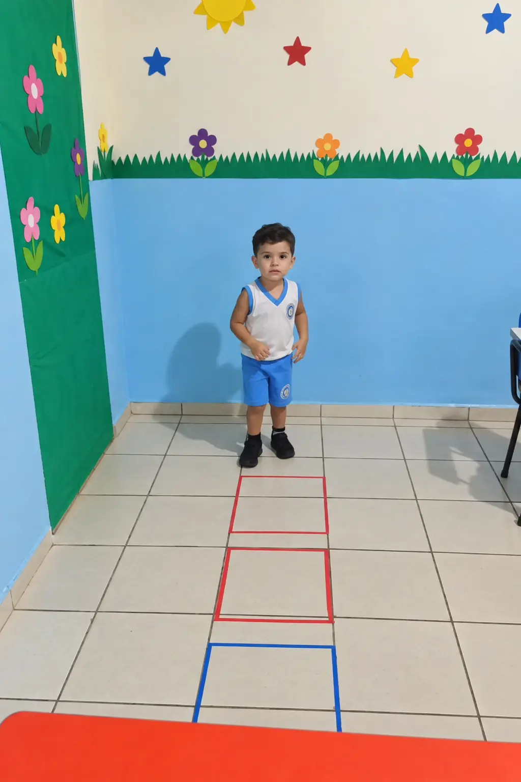 Criança pequena de farda num circuito de amarelinha na sala da Educação Infantil, com parede azul enfeitada de flores e estrelas