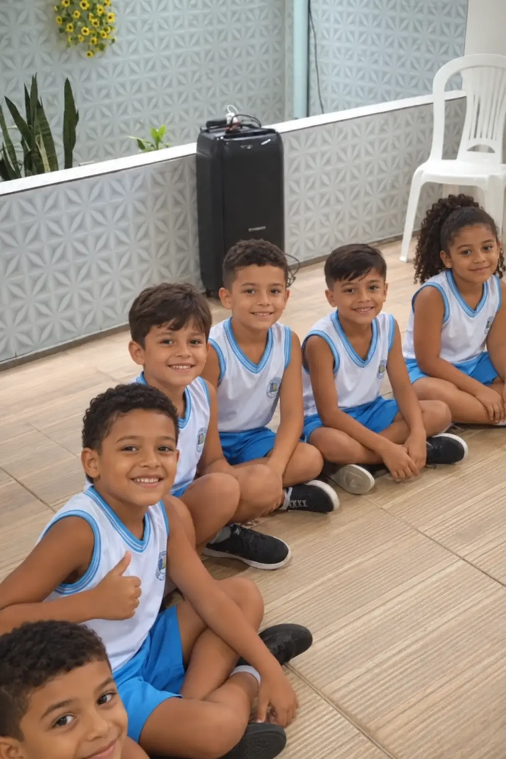 Alunos de farda sentados no chão de uma sala da escola, sorrindo